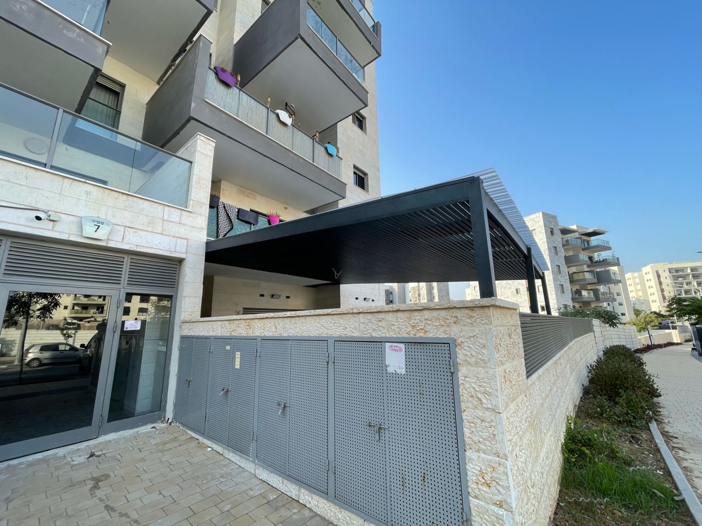 פרגולת אלומיניום כהה בחצר בית לצד בנייני מגורים בחריש