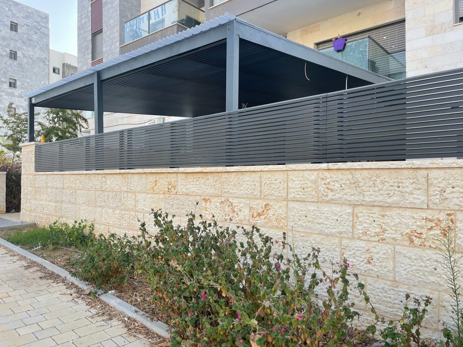 פרגולת אלומיניום כהה עם מסתור רפפות לצד קיר אבן בחריש