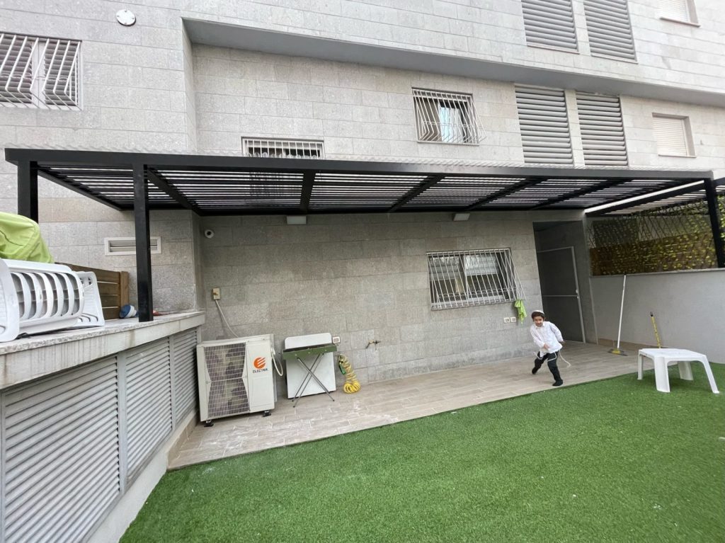 פרגולת אלומיניום כהה מעל חצר עם דשא וקיר ירוק בנתניה